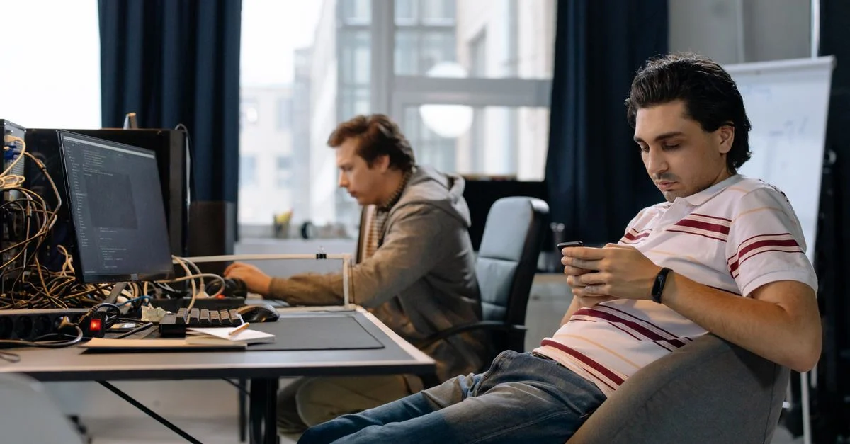 Deux hommes travaillant dans un bureau moderne, l'un sur un ordinateur et l'autre sur un smartphone.