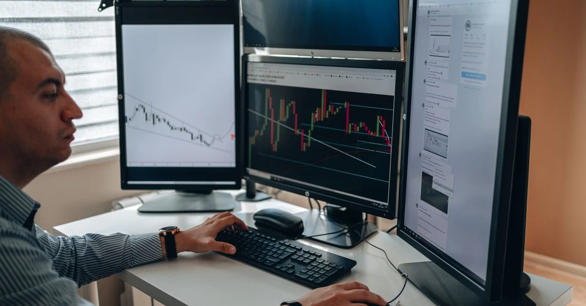 Un professionnel dans un bureau analysant des graphiques financiers sur plusieurs moniteurs, utilisant une technologie avancée.